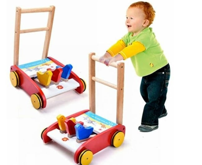 Bé gái tập đi với xe tập đi gỗ dạng đẩy hình chữ L, một lựa chọn an toàn hơn khi cha mẹ cân nhắc có nên cho bé ngồi xe tập đi sớm.